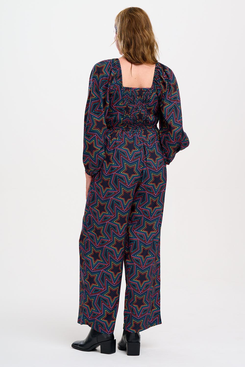 A model is standing and smiling, wearing a black and rainbow star explosion patterned jumpsuit with a square neckline, long blouson sleeves, a fitted waist, and wide-leg trousers.