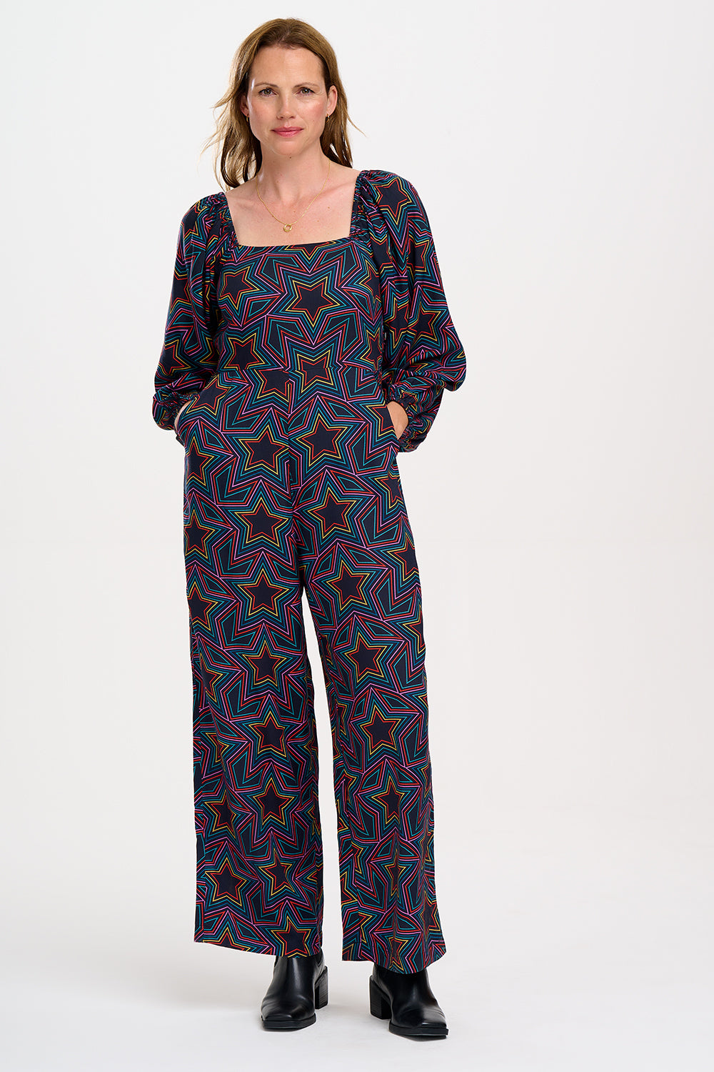 A model is standing and smiling, wearing a black and rainbow star explosion patterned jumpsuit with a square neckline, long blouson sleeves, a fitted waist, and wide-leg trousers.