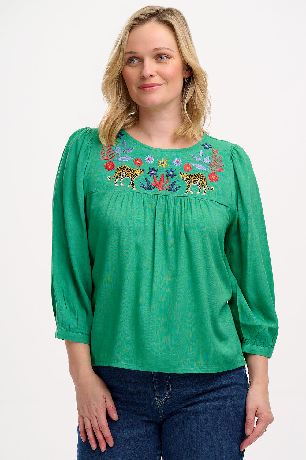A woman wearing a green top with leopard floral embroidery, featuring a relaxed fit with blouson sleeves and gathered detailing, paired with jeans.