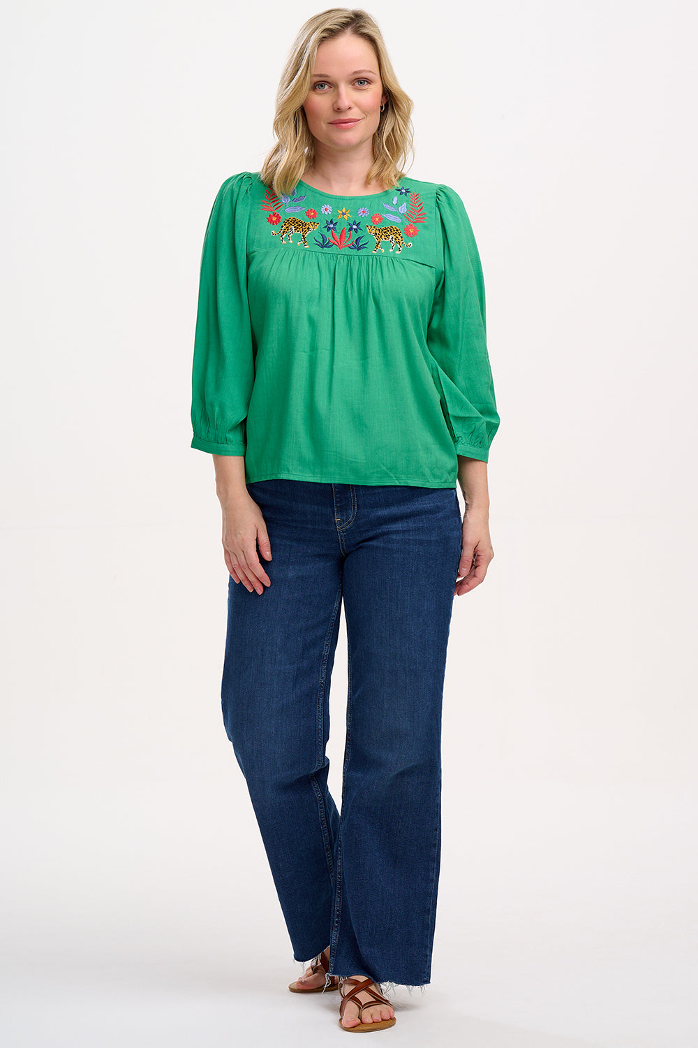 A woman wearing a green top with leopard floral embroidery, featuring a relaxed fit with blouson sleeves and gathered detailing, paired with jeans.