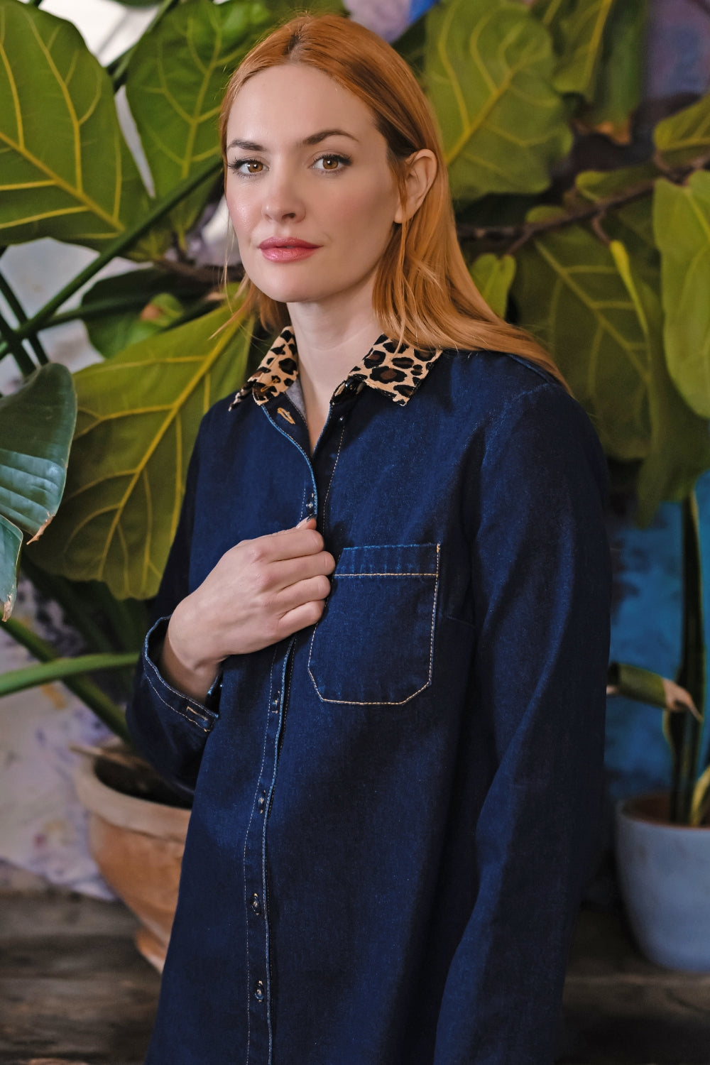 A woman with red hair wearing a dark blue denim shirt-jacket with a leopard-print collar, posed in front of large green potted plants.
