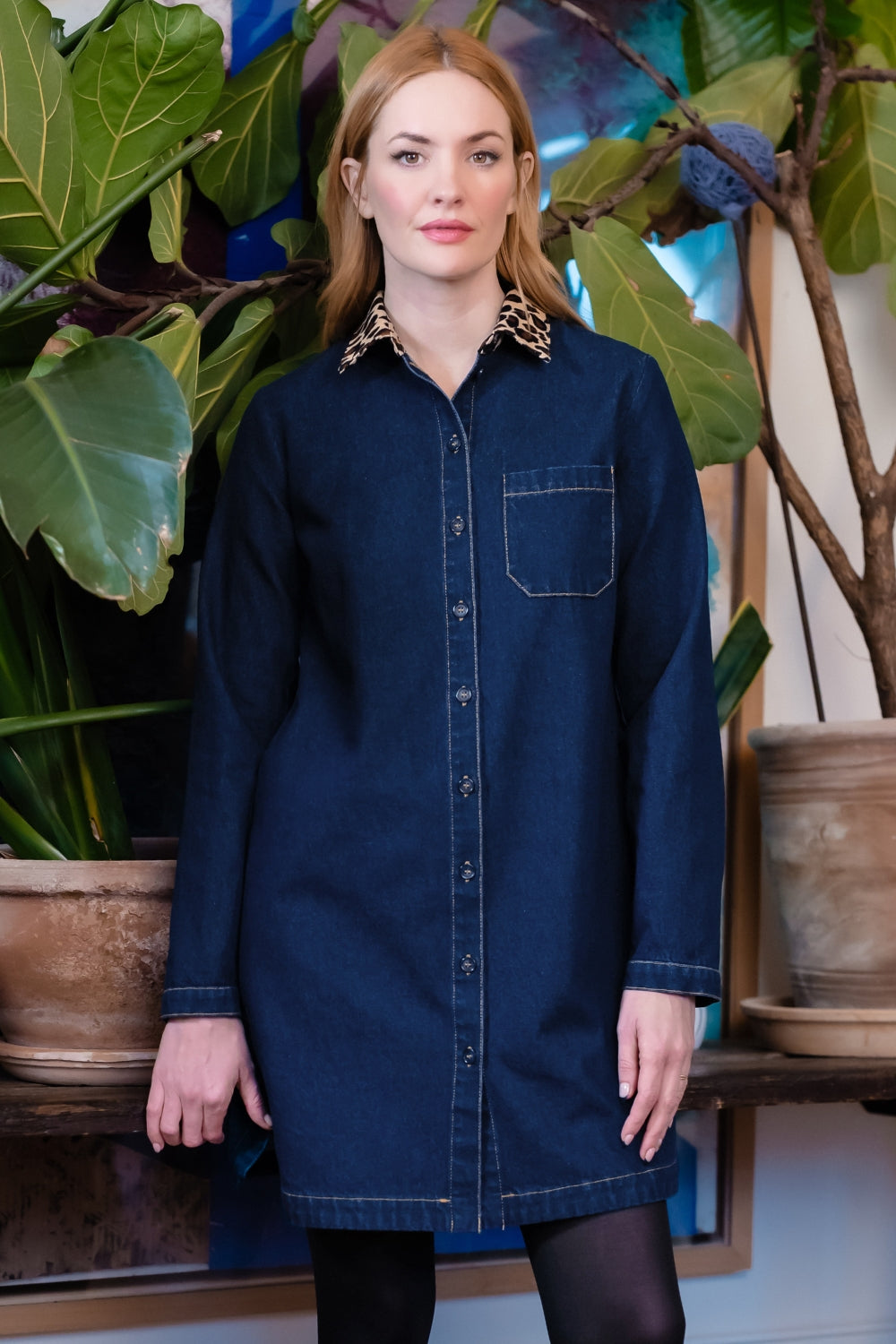 A woman wearing a dark blue denim shirt dress with leopard-print collar, standing in front of a potted plant.