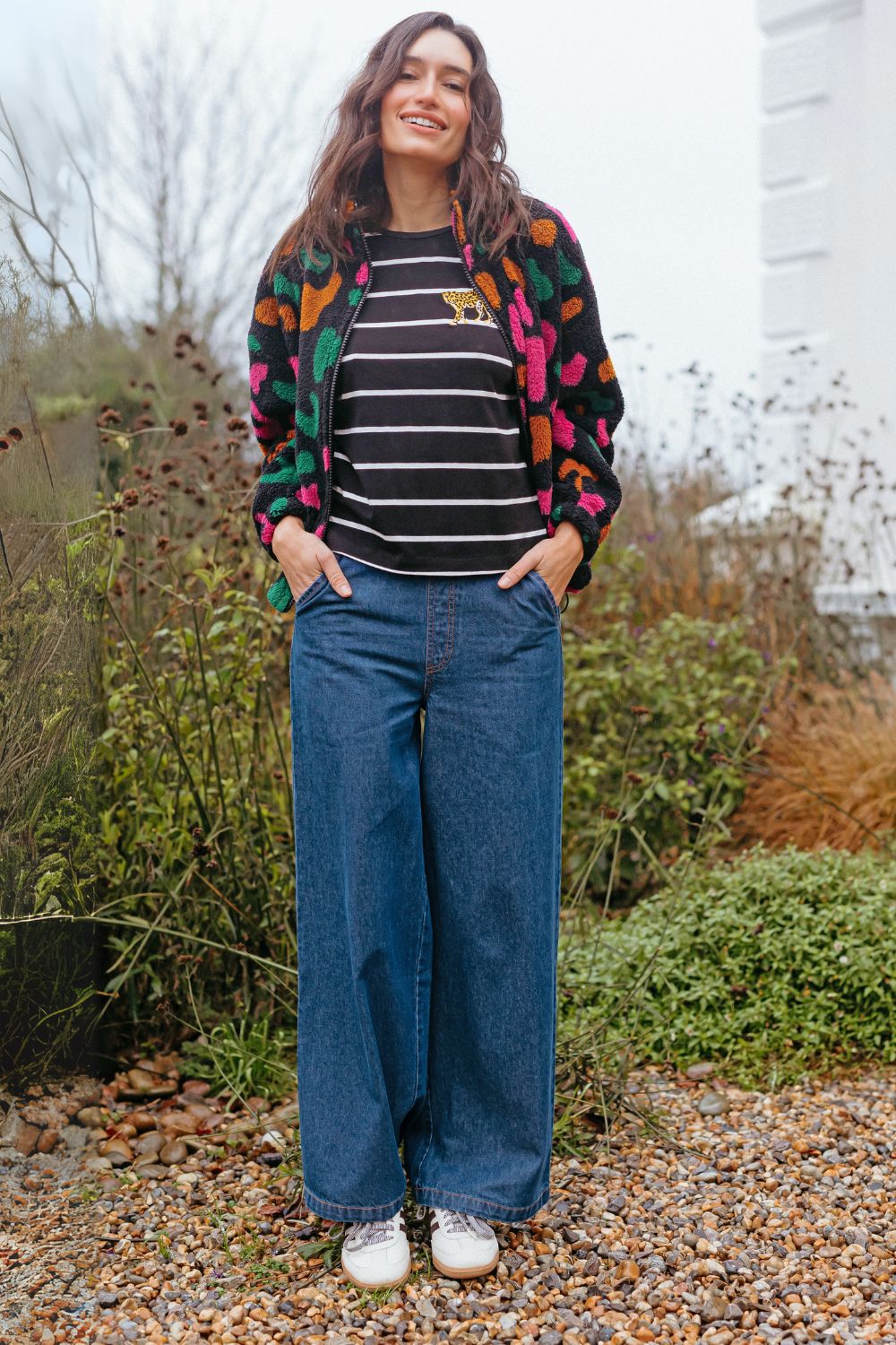 Sugarhill Brighton model wearing a colorful jacket, striped shirt, and blue jeans standing outdoors.