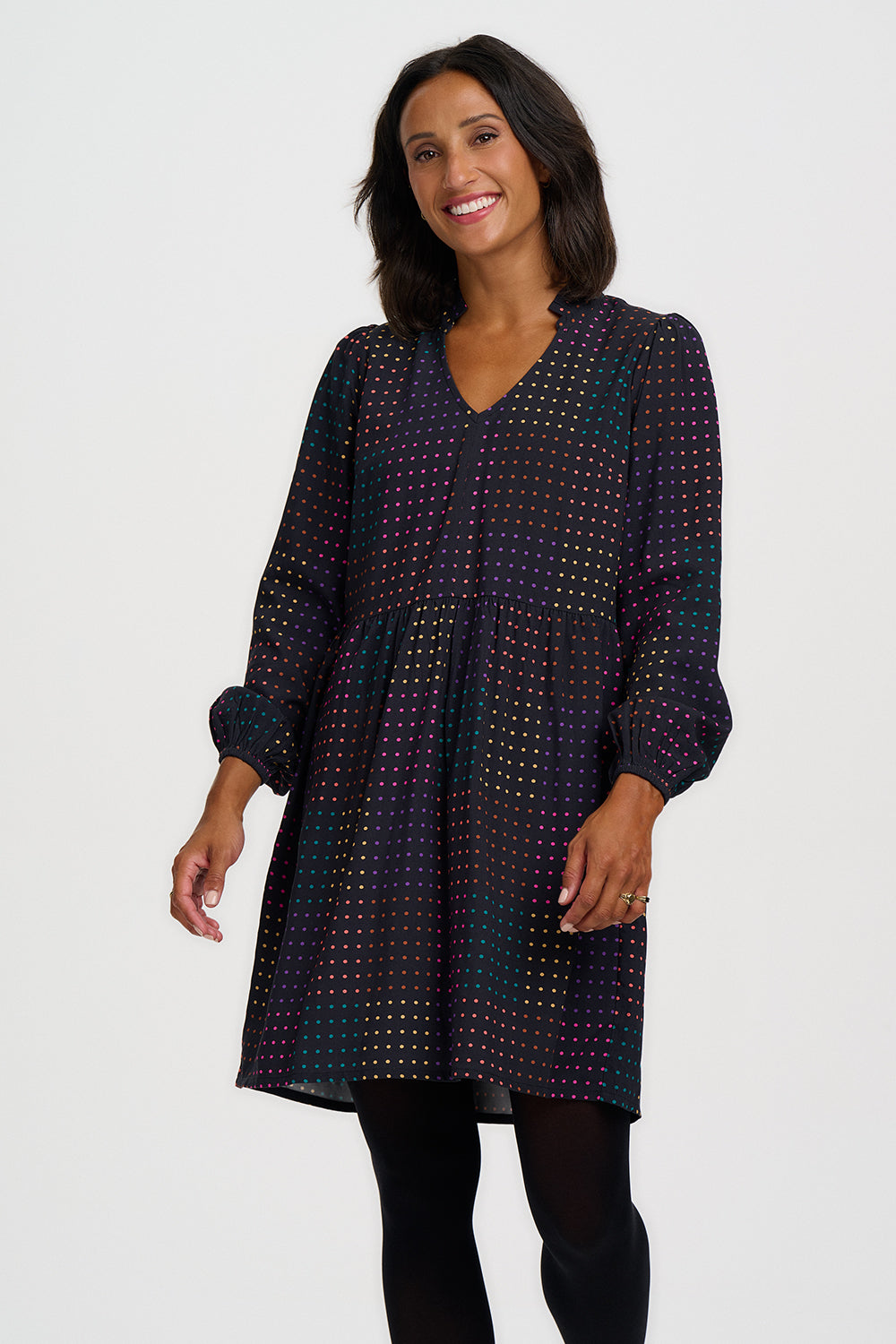 A woman modeling a long-sleeve V-neck dress with a dark base and a dense, small-scale multicolor geometric pattern, paired with black tights against a light gray studio background.