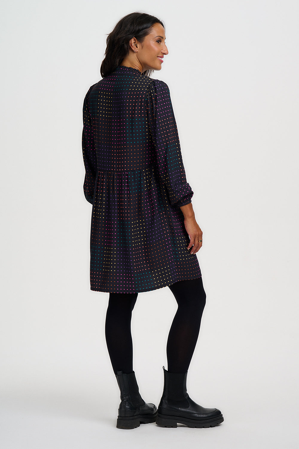 A woman modeling a long-sleeved, knee-length dress with a dark multicolor geometric pattern, paired with black tights and black ankle boots, shown from a side-back angle against a plain white studio background.