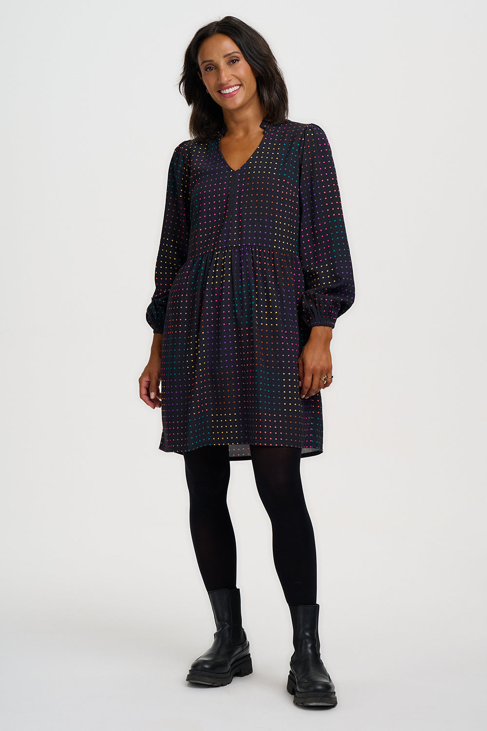 A woman modeling a long-sleeve V-neck mini dress with a dark base and a dense, small-scale multicolor geometric pattern, styled with black tights and black ankle boots against a light gray studio background.