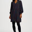 A woman modeling a long-sleeve V-neck mini dress with a dark base and a dense, small-scale multicolor geometric pattern, styled with black tights and black ankle boots against a light gray studio background.