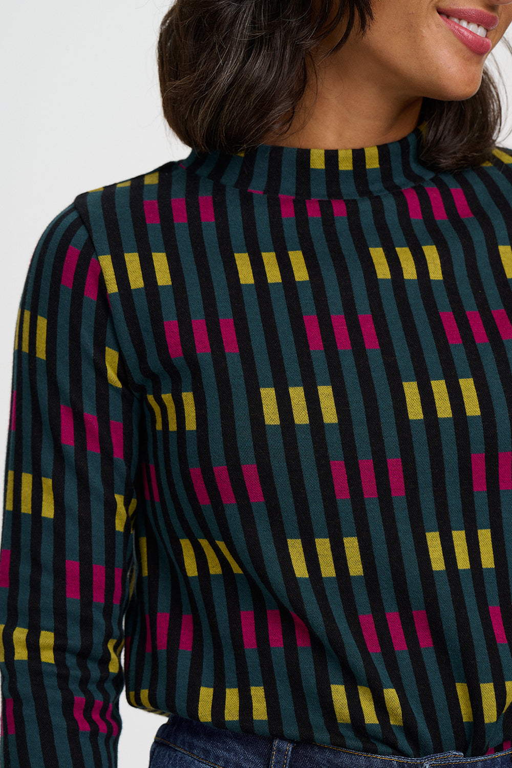 A close-up of a woman wearing a long-sleeve mock neck top with a dark green base and a geometric pattern of vertical black stripes and alternating yellow and magenta rectangular blocks.