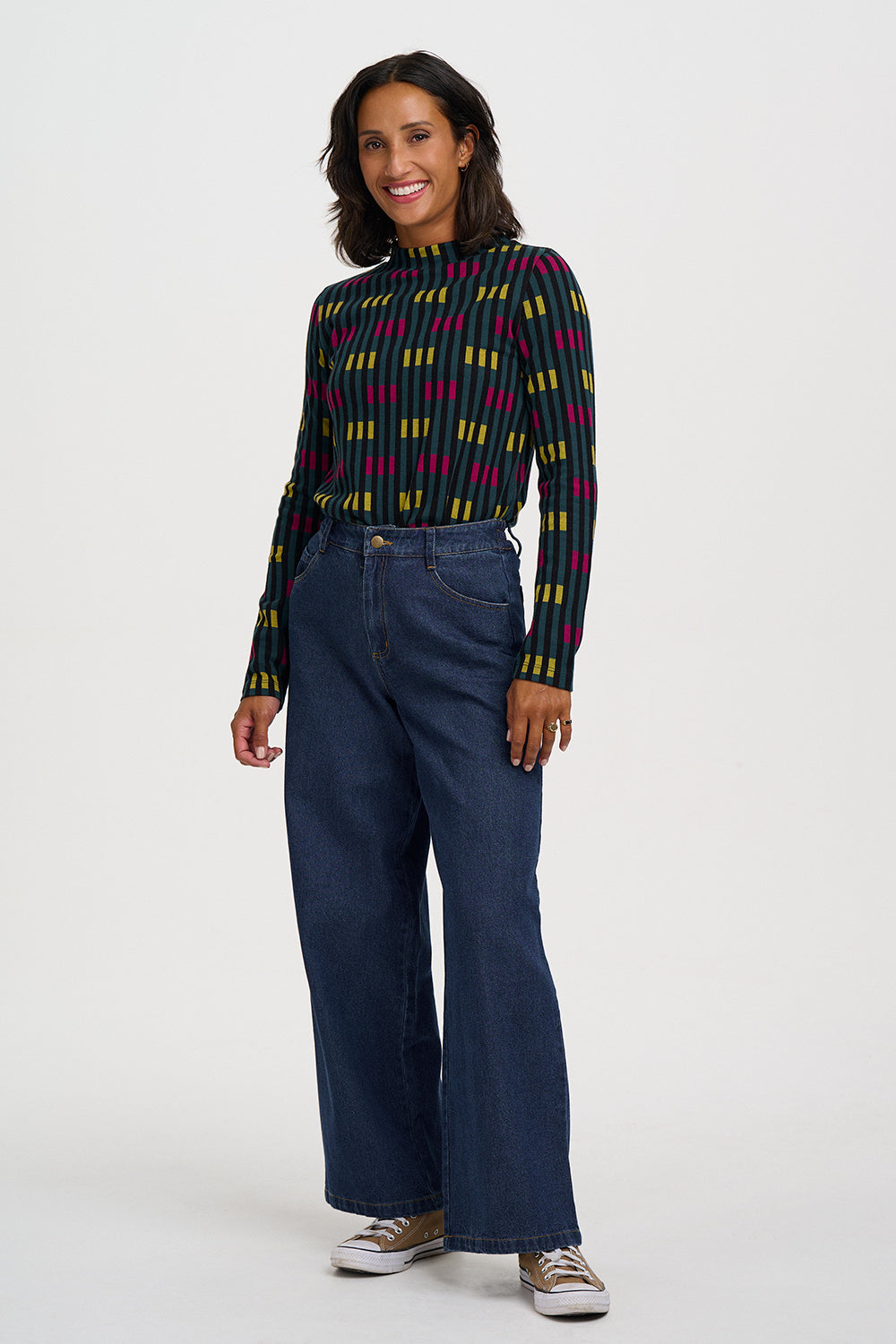 A woman modeling a long-sleeve mock-neck knit top with a multicolor vertical stripe pattern (green, yellow, red, and black) paired with dark blue wide-leg jeans and canvas sneakers, shot against a plain light gray studio background.