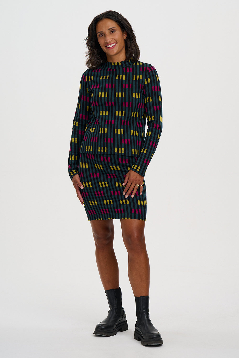 A woman modeling a long-sleeve mini dress with a colorful geometric pattern of yellow, magenta, and light blue lines on a black background, paired with black ankle boots, against a plain light gray studio background.