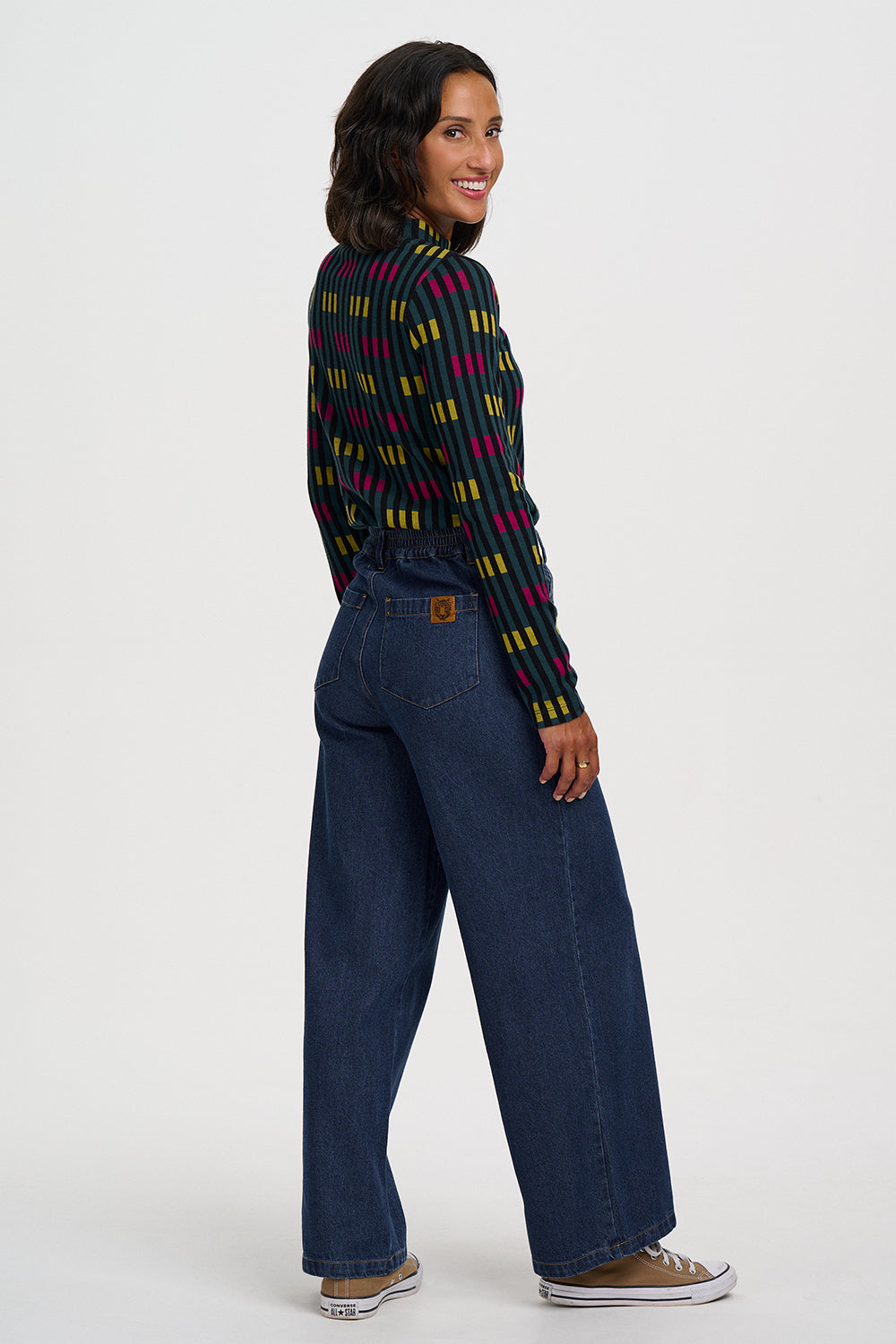 A woman modeling a dark navy wide-leg denim pant with a colorful striped knit top, shown from a three-quarters back view against a plain white studio background.