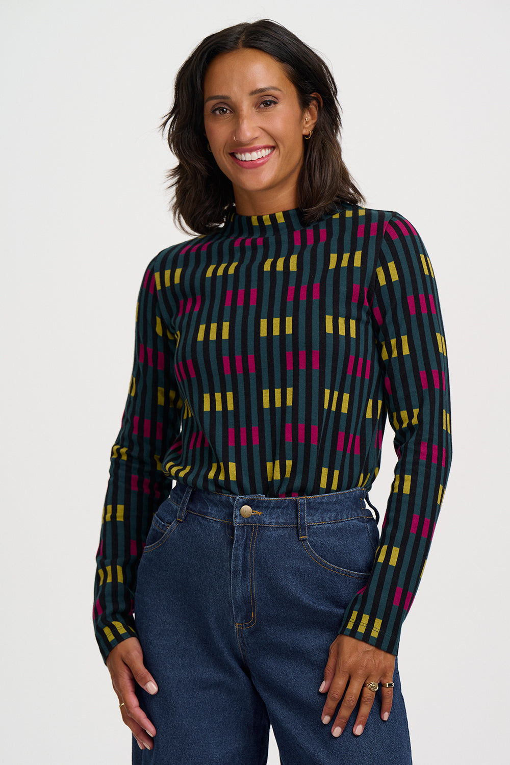 A woman wearing a long-sleeve mock neck top with a dark green base and a geometric pattern of yellow and magenta vertical stripes, paired with high-waisted blue jeans, against a light gray studio background.
