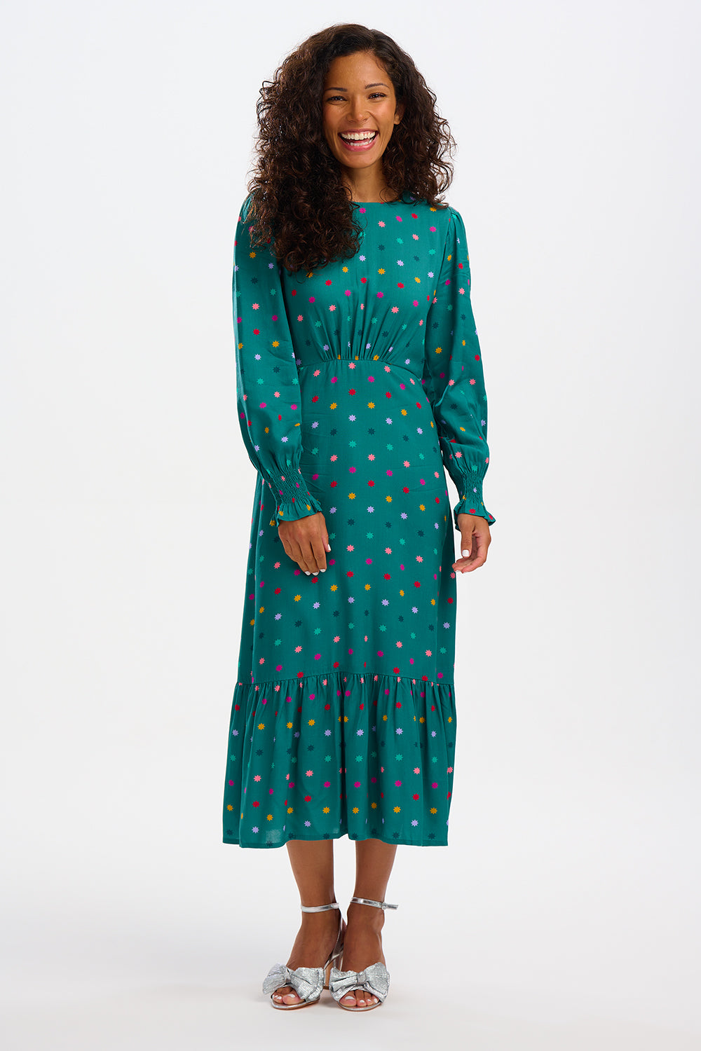 A woman standing and smiling, wearing a green maxi dress with a winter starburst pattern and long sleeves.