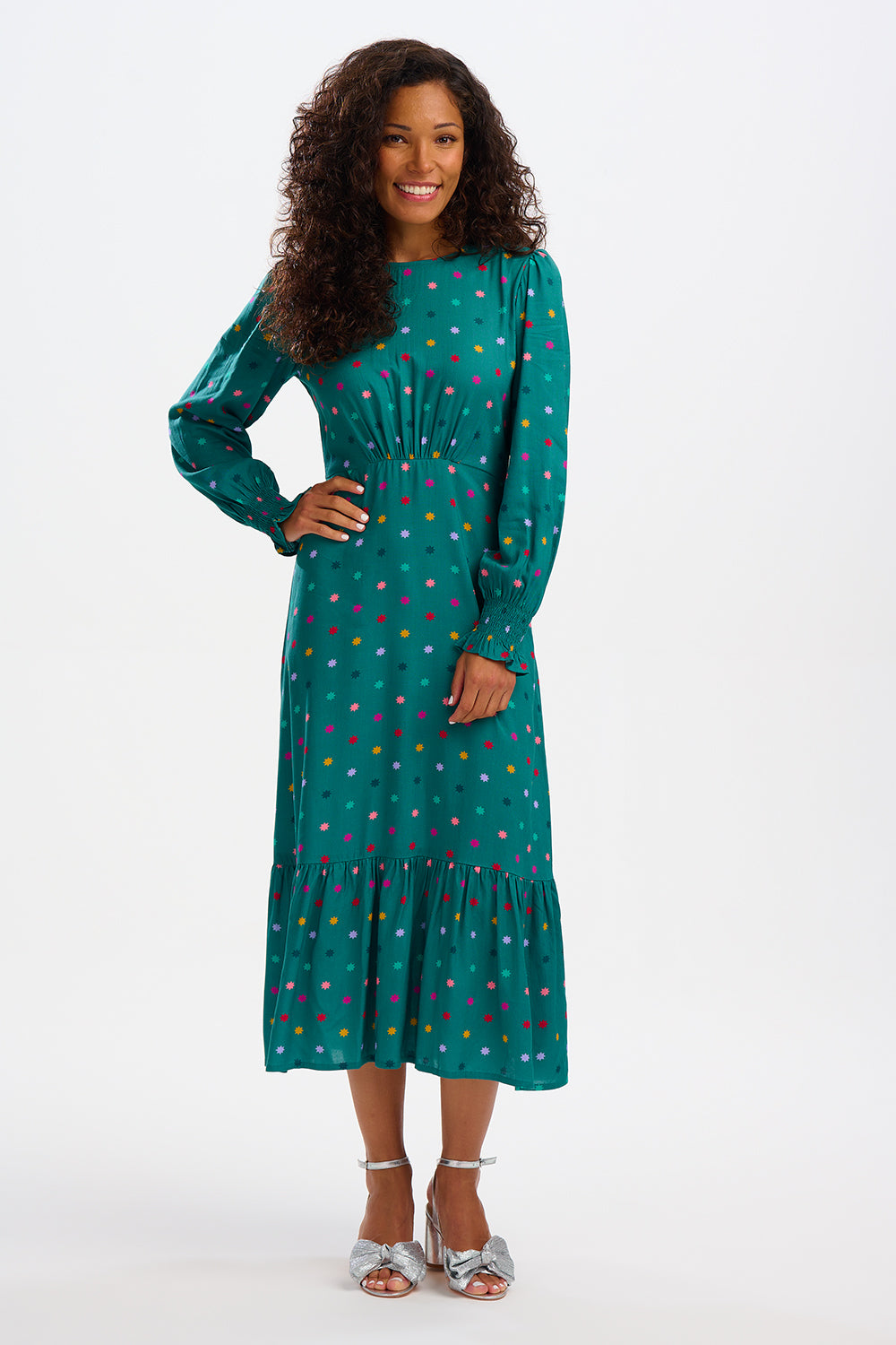 A woman standing and smiling, wearing a green maxi dress with a winter starburst pattern and long sleeves.