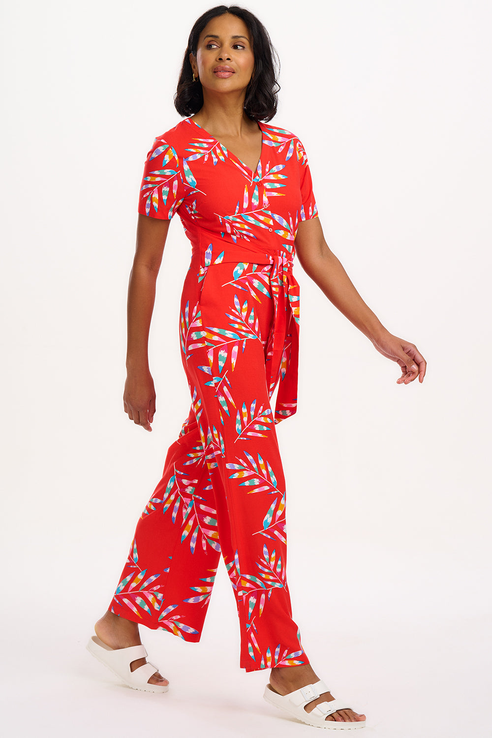 A model wearing a red jumpsuit with a tropical leaf print, featuring a cap-sleeved design, wide legs, and a self-fabric belt at the waist.