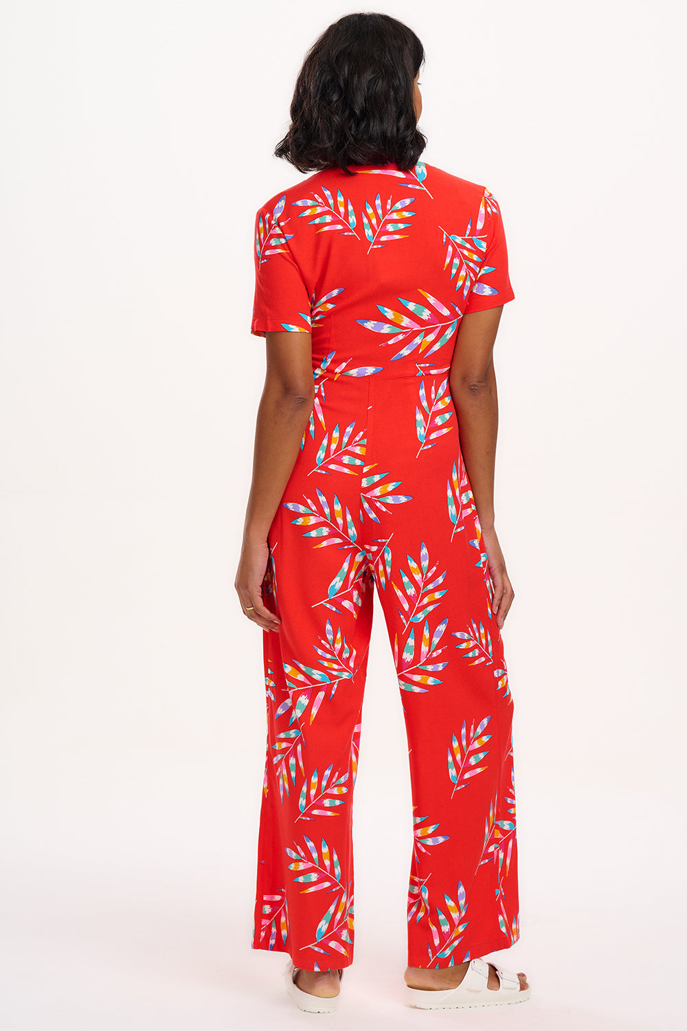 A red jumpsuit with a rainbow frond pattern, featuring a round neckline and a tie-waist belt.