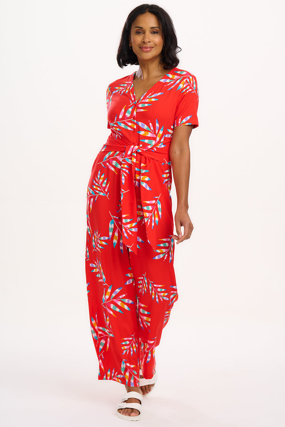 A red jumpsuit with a rainbow frond pattern, featuring a round neckline and a tie-waist belt.