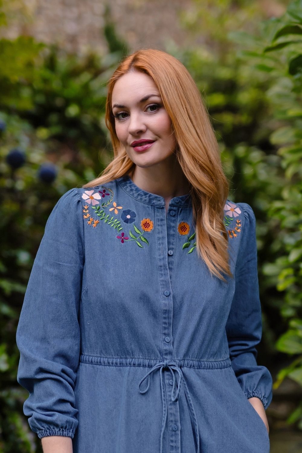 A woman with long red hair wearing a blue denim-style long-sleeve shirt dress with colorful floral embroidery on the chest, standing outdoors in front of a blurred green garden background.
