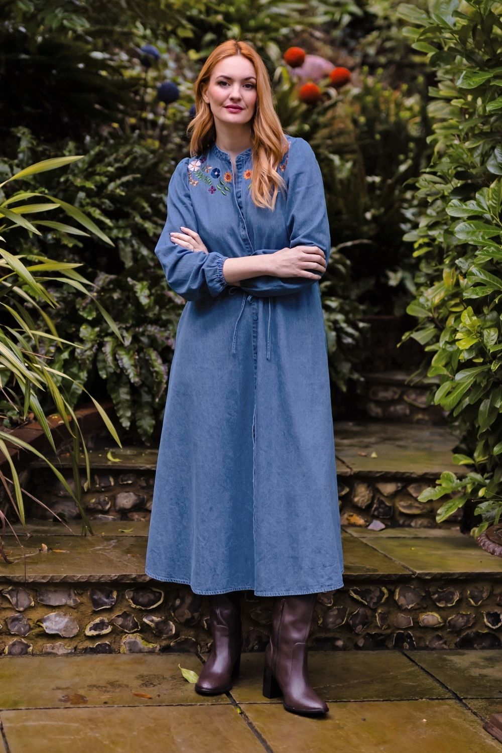 A woman with red hair stands in a garden wearing a long-sleeved blue midi dress with colorful embroidered details on the chest and brown leather boots.