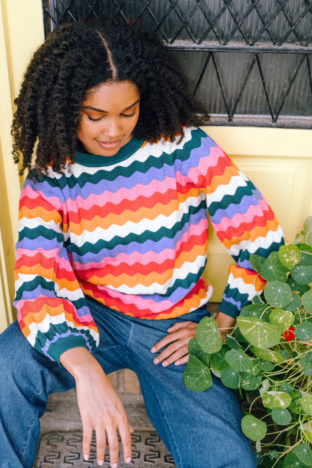 A woman sitting beside a plant, wearing a multicolored striped jumper with a round neckline and long sleeves.