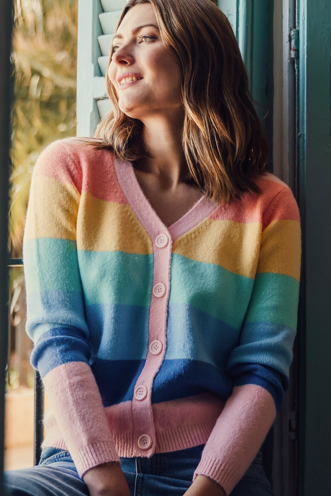 A woman wearing a multicolored striped cardigan with a deep v neckline and large buttons, in pastel colors such as pink, yellow, blue, and green.