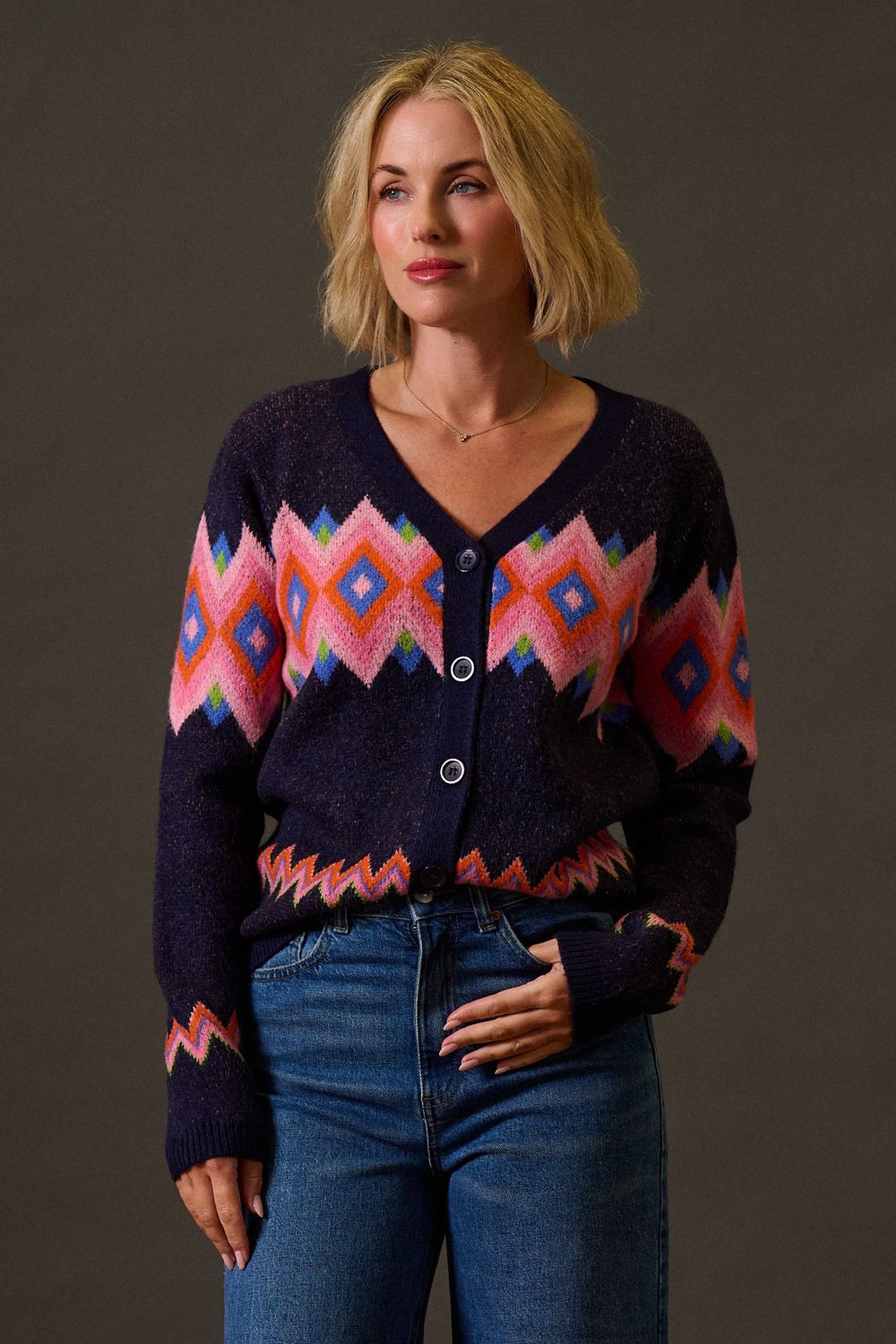 Woman wearing a dark blue cardigan with colorful geometric patterns and blue jeans against a brown background