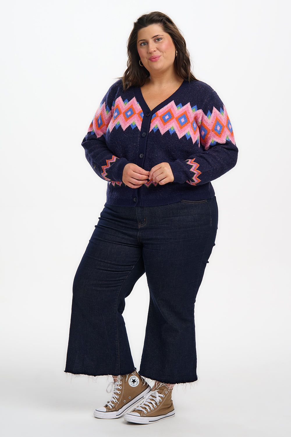 Woman wearing a navy cardigan with pink and blue pattern, dark jeans, and brown sneakers on a white background