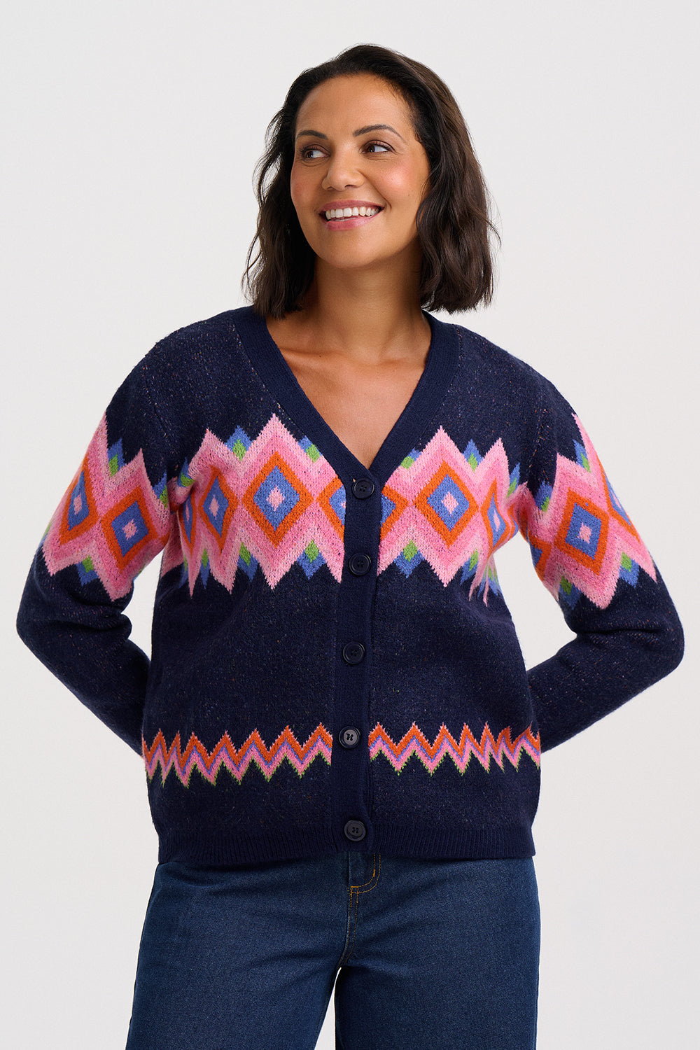 A woman wearing a navy blue button-up cardigan with a colorful geometric pattern in pink, orange, and blue, paired with dark blue jeans, against a light gray studio background.