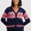 A woman wearing a navy blue button-up cardigan with a colorful geometric pattern in pink, orange, and blue, paired with dark blue jeans, against a light gray studio background.