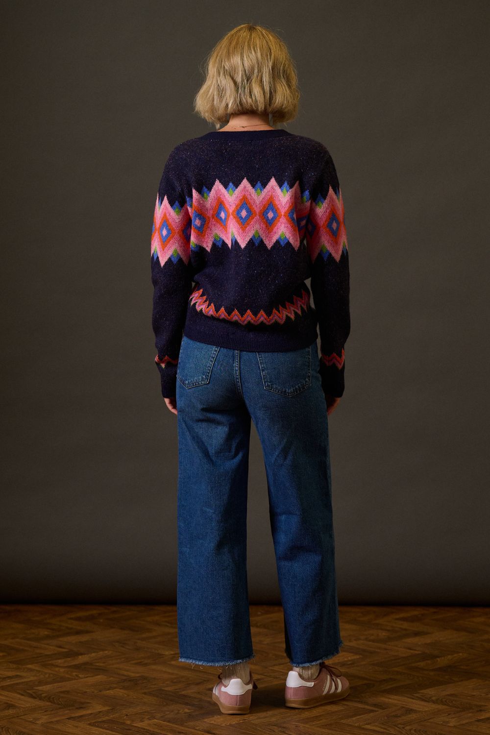 Person wearing a patterned sweater and blue jeans against a dark background
