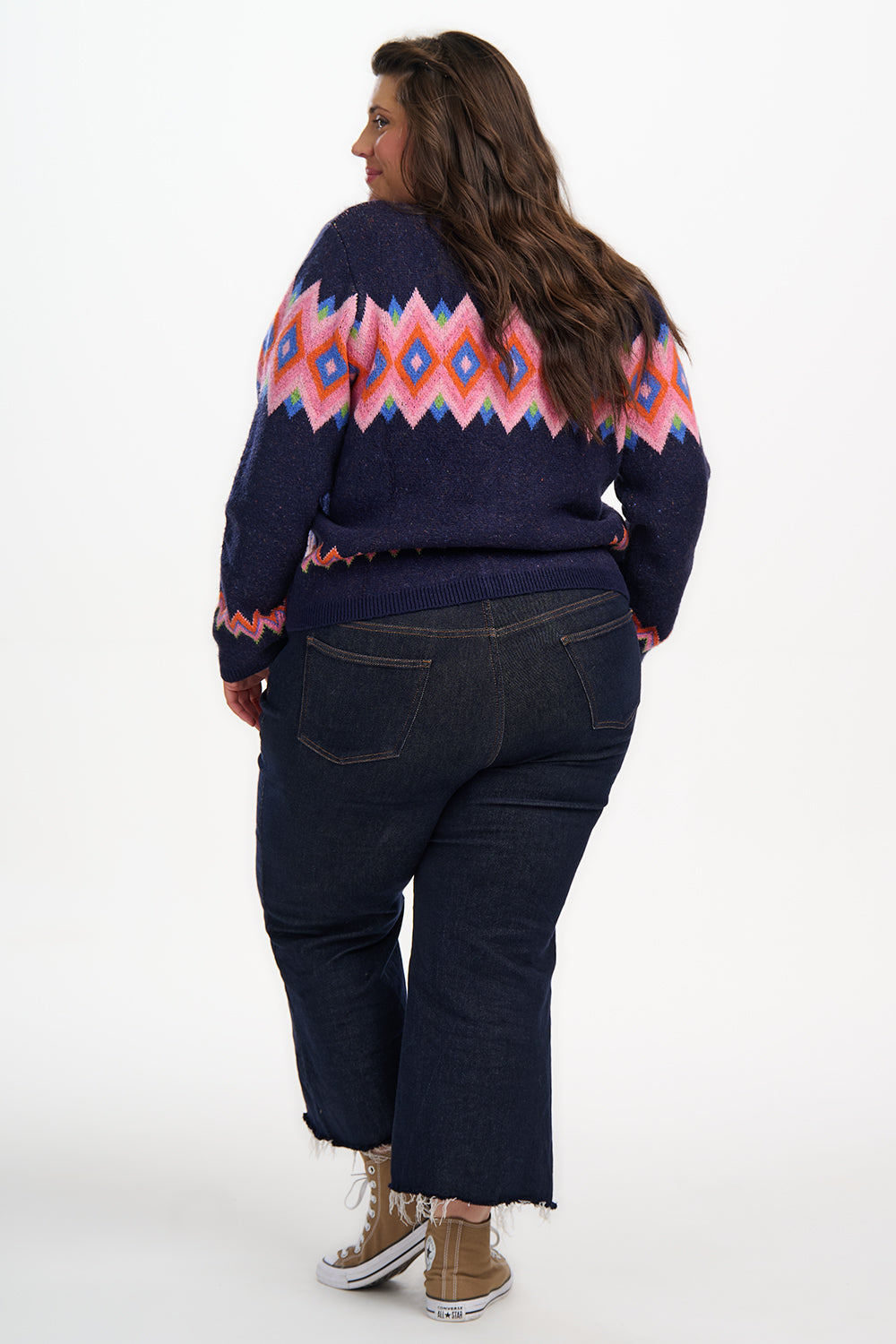 Person wearing a navy sweater with a colorful pattern and dark jeans on a white background
