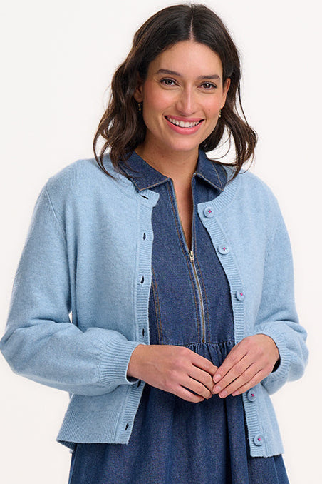 A woman smiling, wearing a sky blue knit cardigan with a round neck, buttoned front, and blouson sleeves, paired with a denim dress.