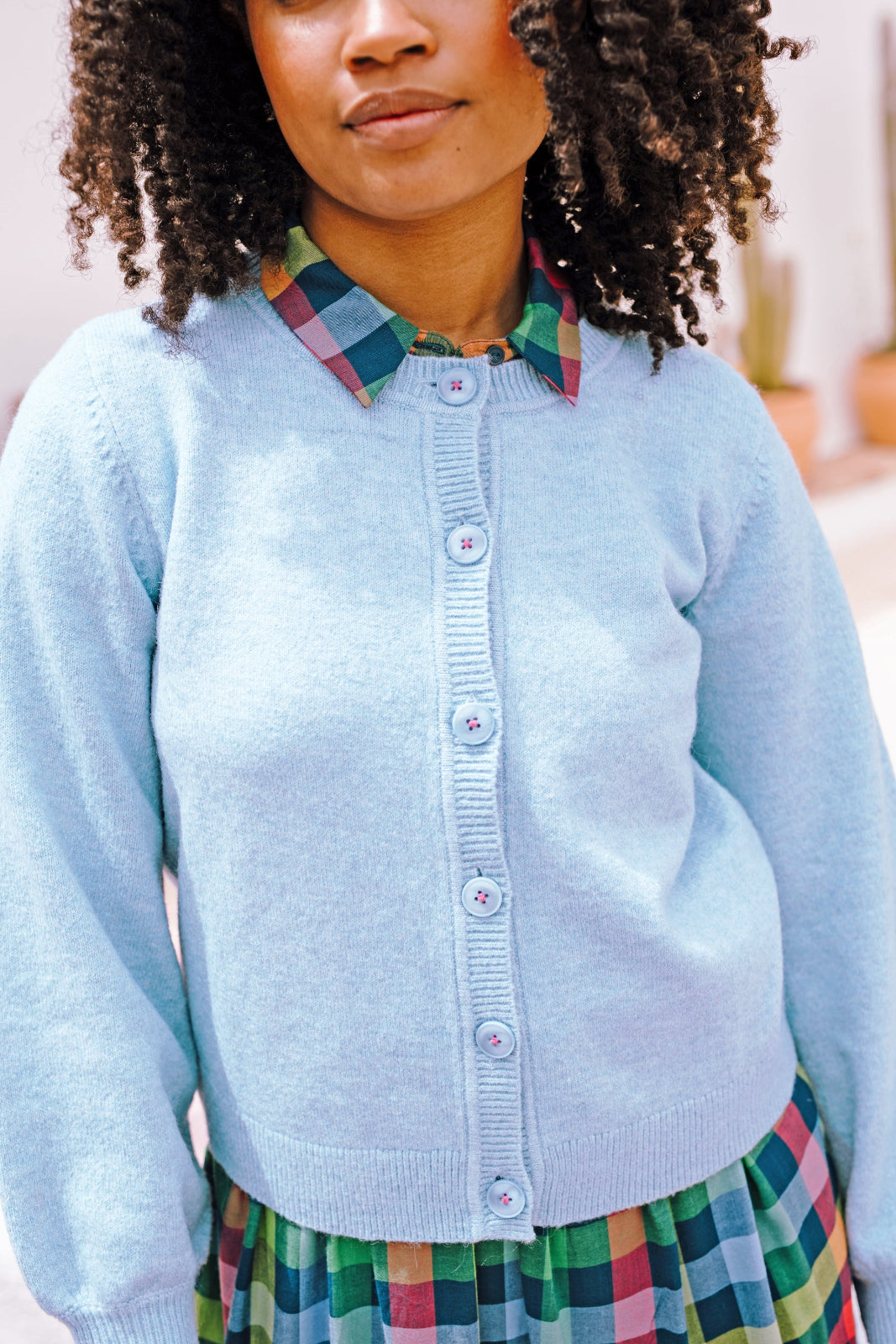 A woman wearing a sky blue Jamie Cardigan with ribbed round neck, buttoned front, and blouson sleeve. The cardigan features small matching sky blue buttons with accent hot pink stitching.
