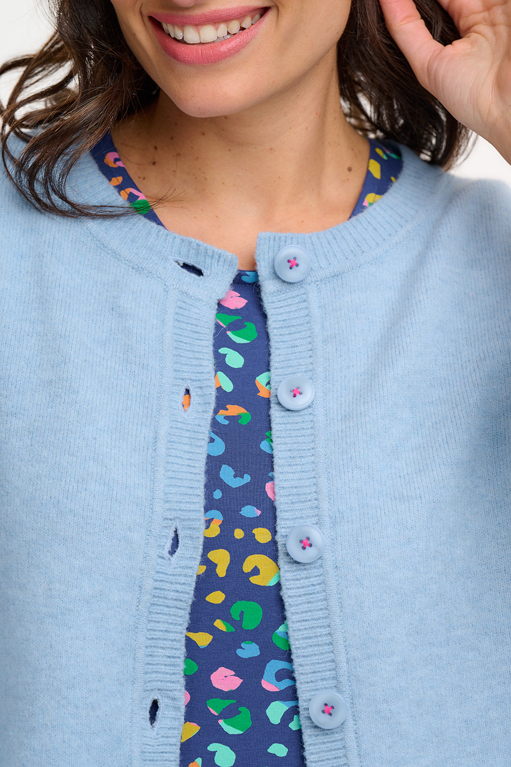 A woman wearing a sky blue Jamie Cardigan with ribbed round neck, buttoned front, and blouson sleeve. The cardigan features small matching sky blue buttons with accent hot pink stitching.