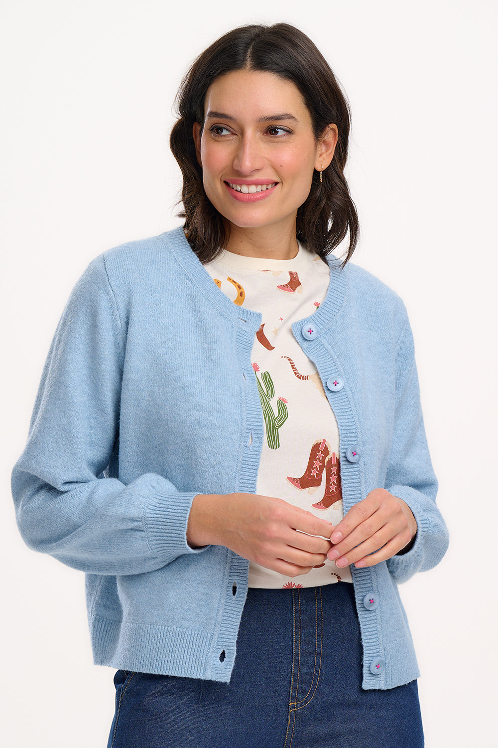 A woman wearing a sky blue Jamie Cardigan with ribbed round neck, buttoned front, and blouson sleeve. The cardigan features small matching sky blue buttons with accent hot pink stitching.