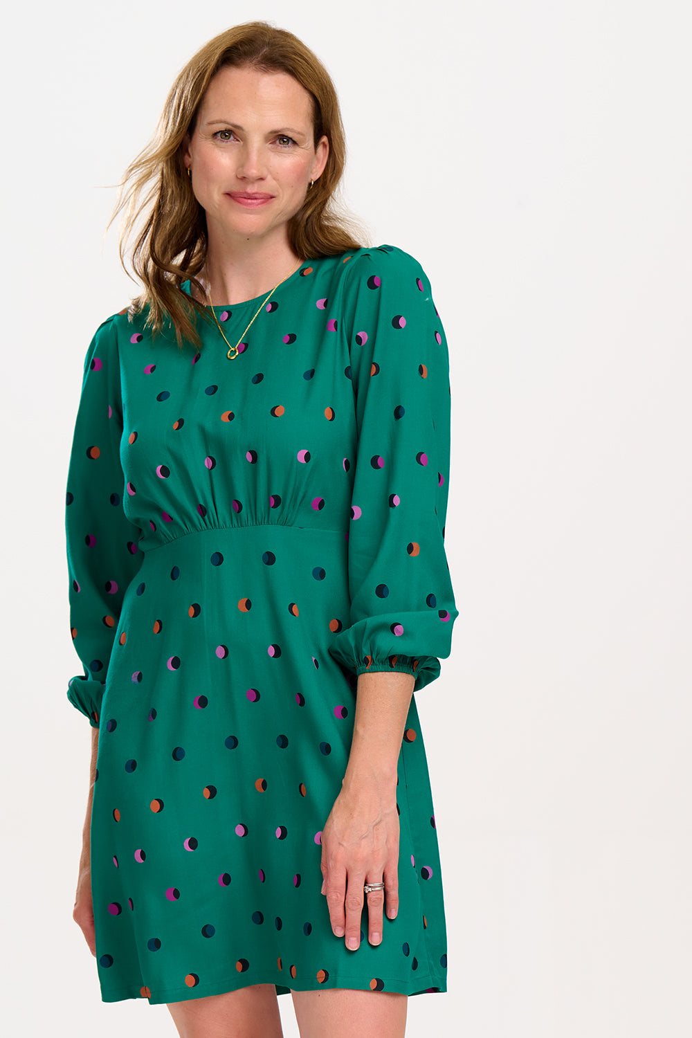 A woman wearing a green mini dress with a rainbow shadow polka print, featuring gathers at the shoulders, long blouson sleeves, a fitted waist, and a flared skirt.