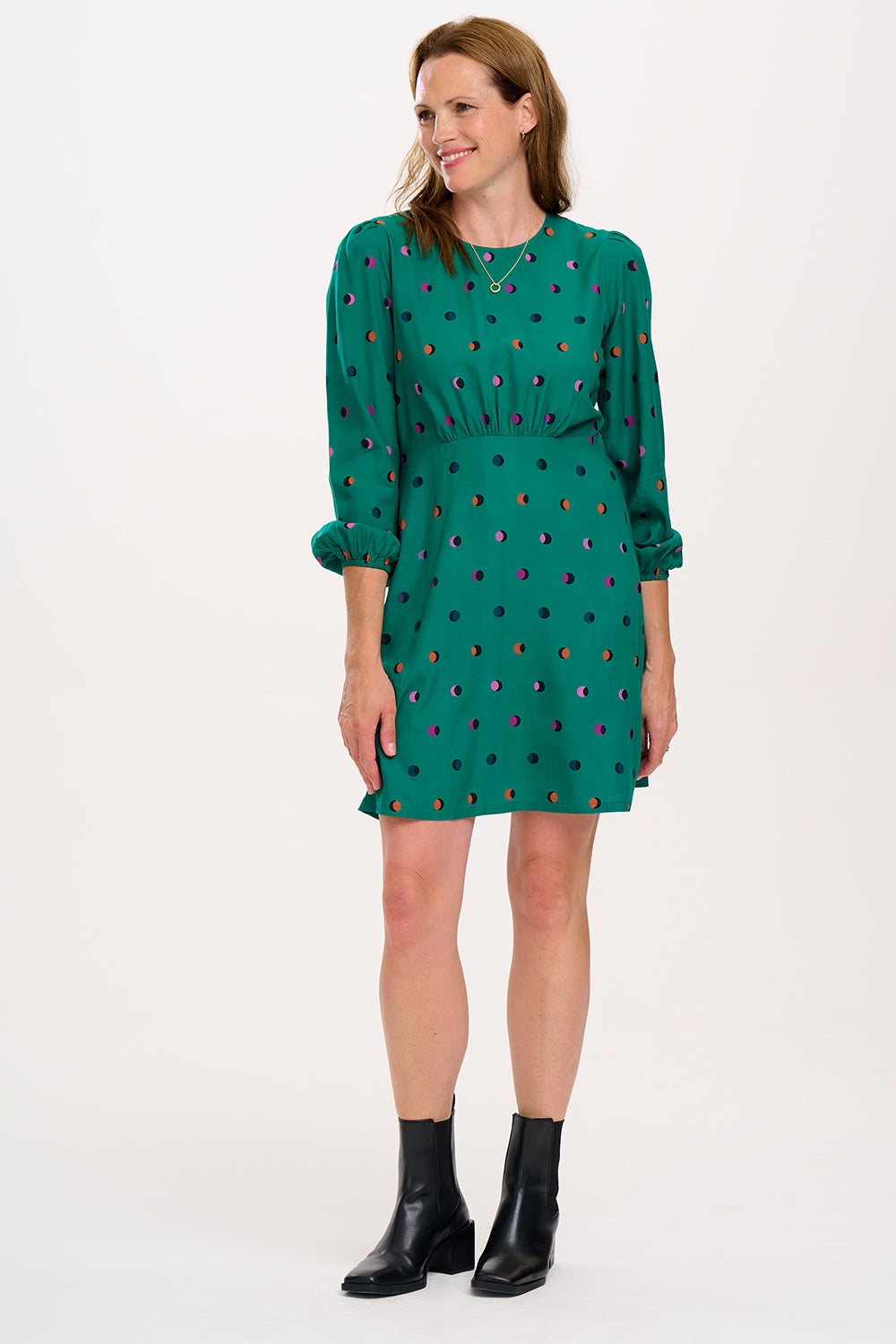 A woman wearing a green mini dress with a rainbow shadow polka print, featuring gathers at the shoulders, long blouson sleeves, a fitted waist, and a flared skirt.