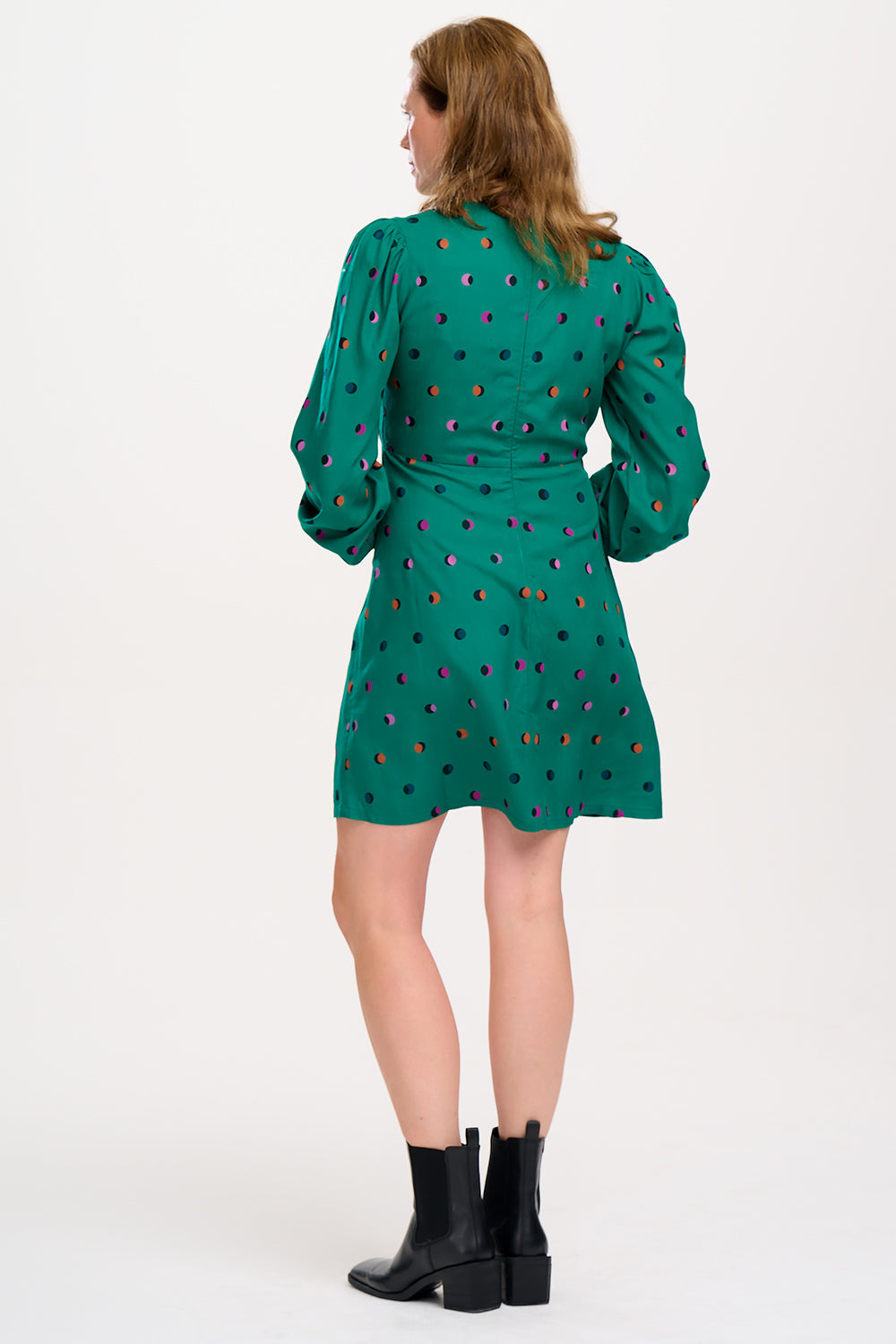 A woman wearing a green mini dress with a rainbow shadow polka print, featuring gathers at the shoulders, long blouson sleeves, a fitted waist, and a flared skirt.
