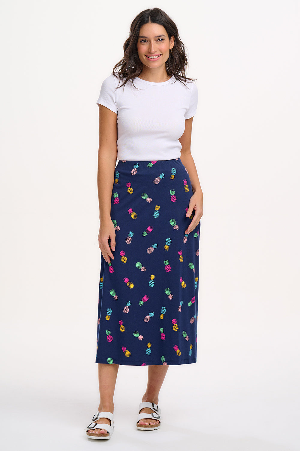 A woman standing and wearing a navy A-line skirt with a multicolor pineapple print, paired with a white top and sandals.