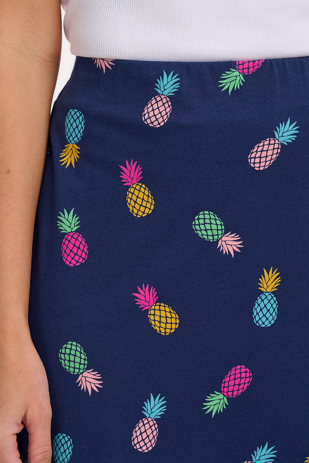 A woman standing and wearing a navy A-line skirt with a multicolor pineapple print, paired with a white top and sandals.