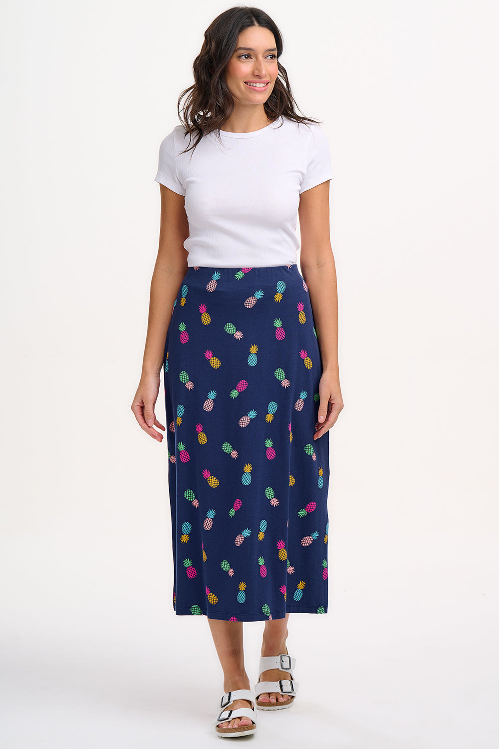 A woman standing and wearing a navy A-line skirt with a multicolor pineapple print, paired with a white top and sandals.