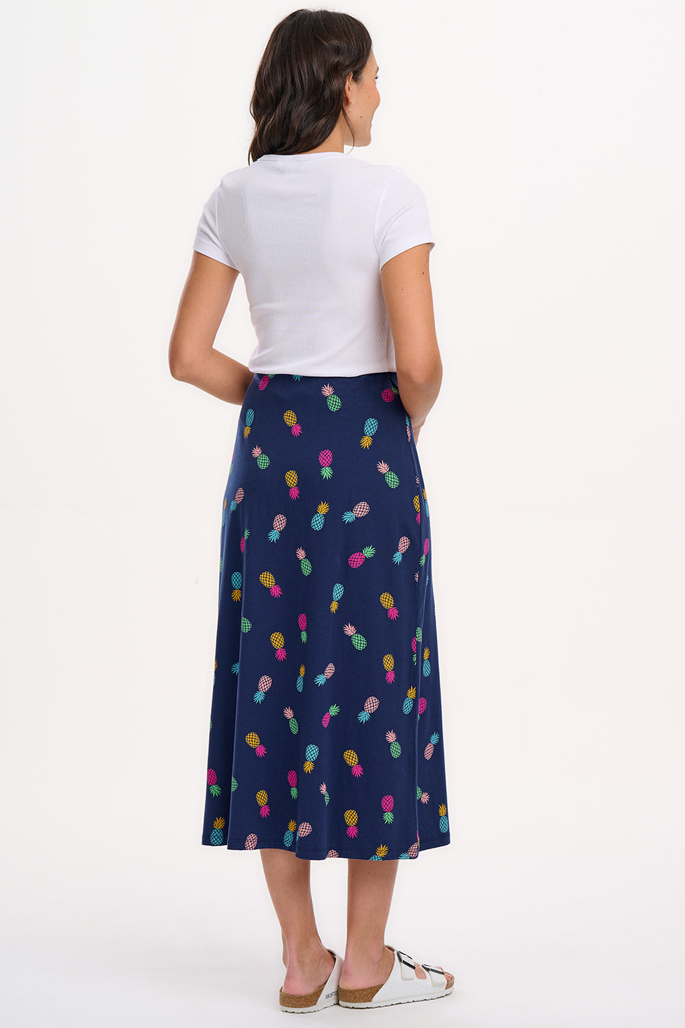 A woman standing and wearing a navy A-line skirt with a multicolor pineapple print, paired with a white top and sandals.