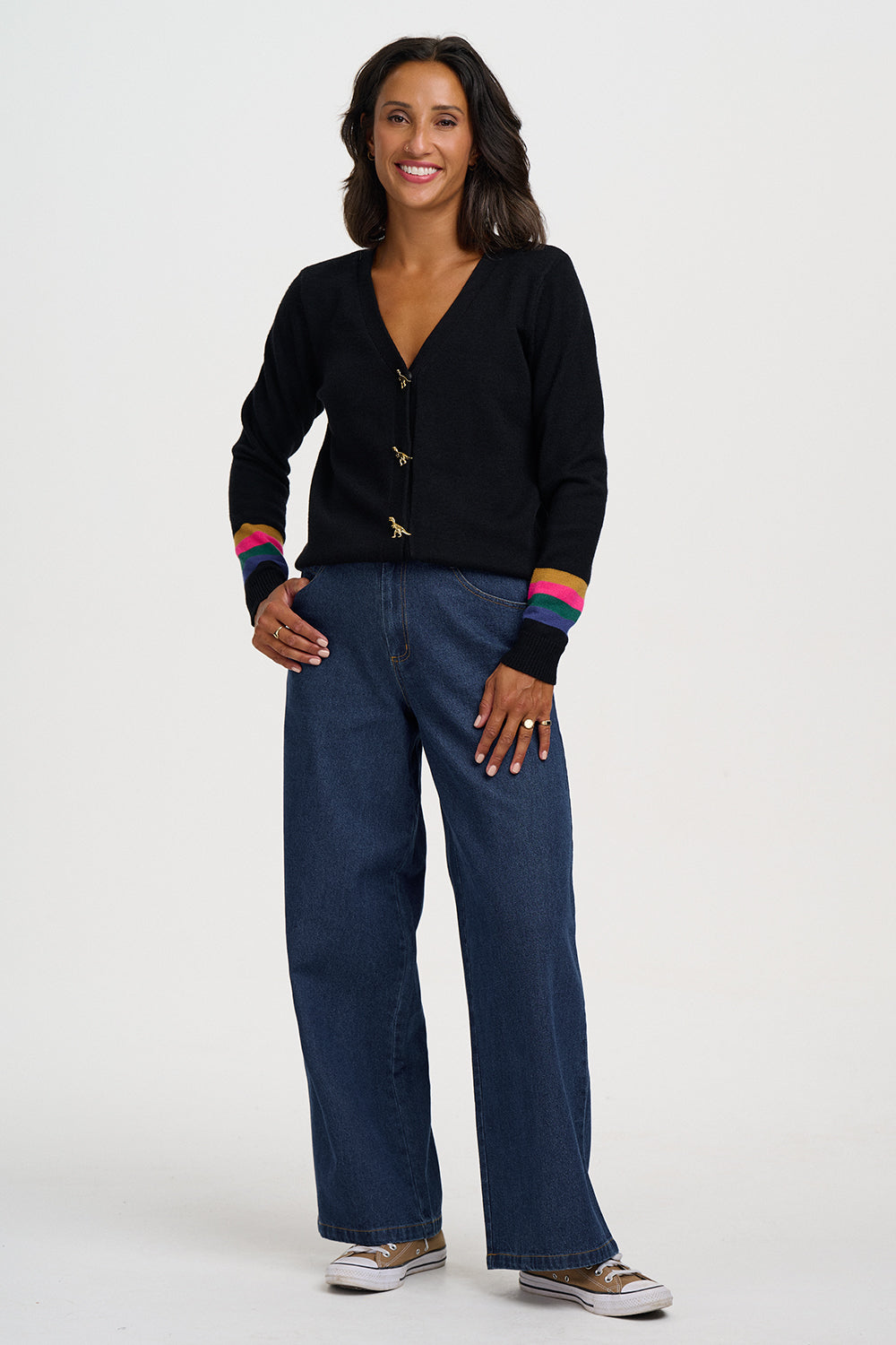 A woman modeling a black long-sleeve cardigan with colorful striped cuffs and gold buttons, paired with dark blue wide-leg jeans and brown sneakers, against a plain light gray studio background.
