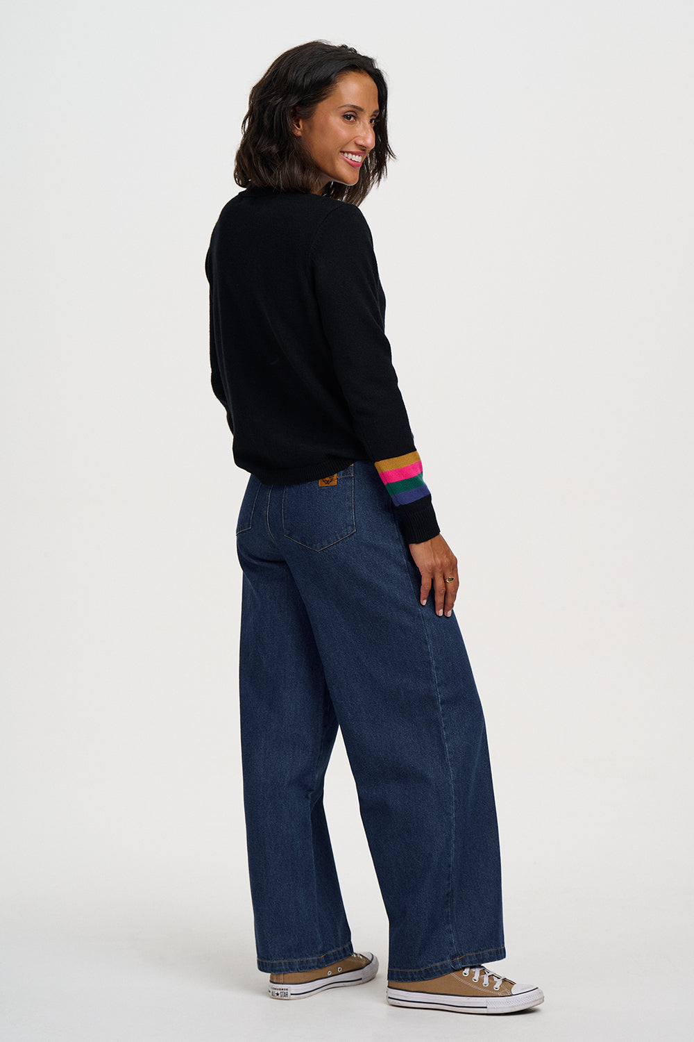 A woman wearing a black long-sleeve top with colorful striped cuffs, dark blue wide-leg jeans, and brown sneakers, shown from a side-back angle against a plain light gray studio background.