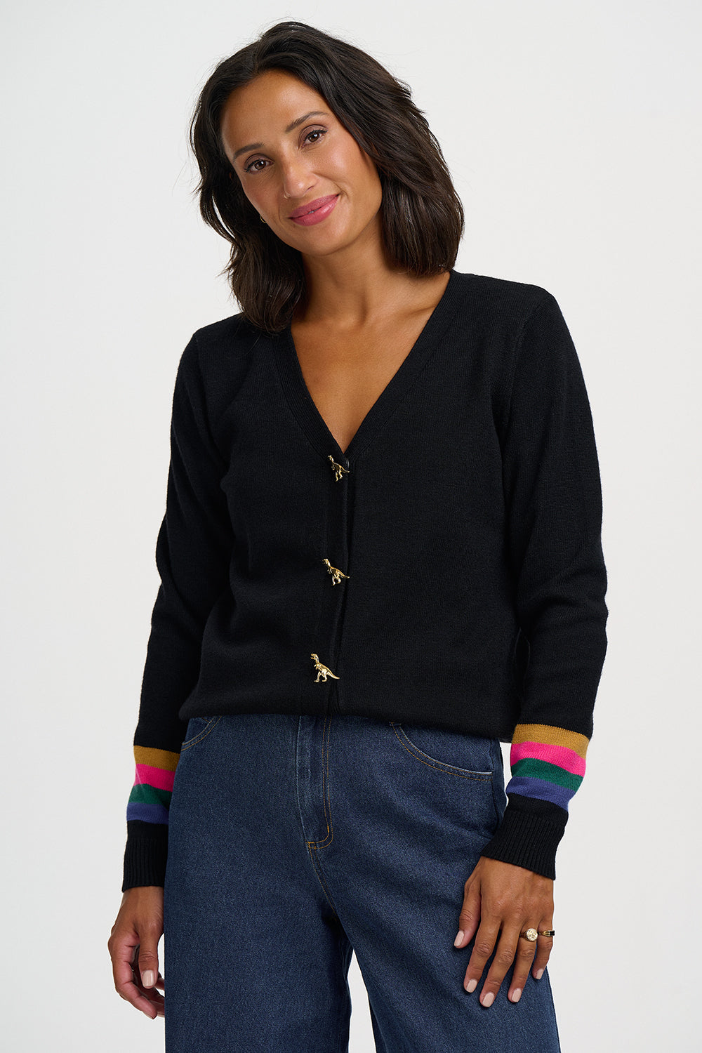 A woman wearing a black knit cardigan with gold-tone bird-shaped buttons and colorful striped cuffs, paired with dark blue jeans, against a plain light gray studio background.