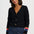 A woman wearing a black knit cardigan with gold-tone bird-shaped buttons and colorful striped cuffs, paired with dark blue jeans, against a plain light gray studio background.