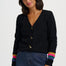 A woman wearing a black knit cardigan with gold-tone bird-shaped buttons and colorful striped cuffs, paired with dark blue jeans, against a plain light gray studio background.