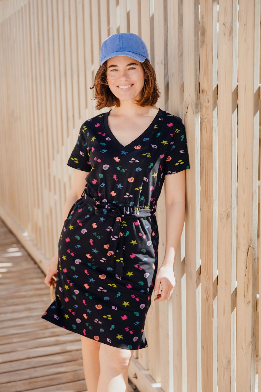 Woman wearing a black dress with colorful patterns and a blue cap, standing against a wooden background.