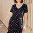 Woman wearing a black dress with colorful patterns and a blue cap, standing against a wooden background.