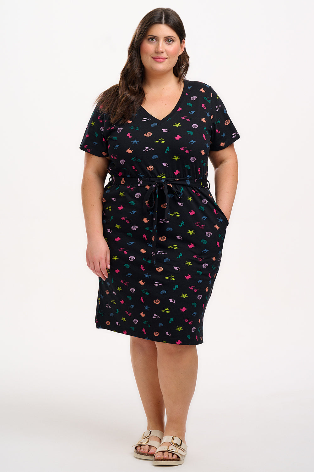 A model wearing a short-sleeved black mini dress with a V-neck, featuring a multicolored underwater theme print. The dress has a matching tie belt and pockets on the skirt.